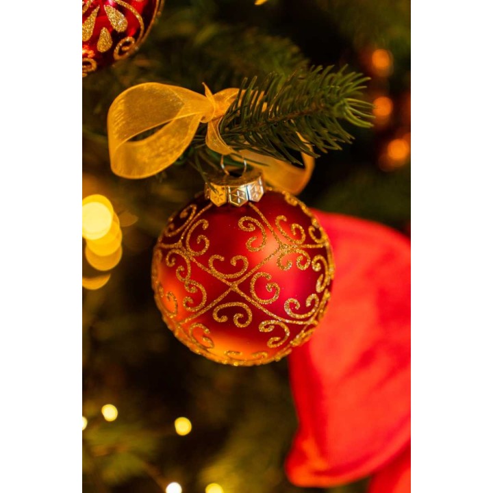 A shiny red ornament with gold swirls hangs from a green pine branch, surrounded by warm lights.