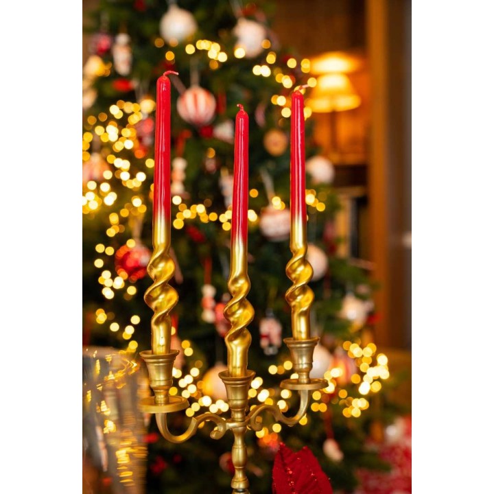 A golden candle holder with three twisted red candles, set against a festive tree backdrop.