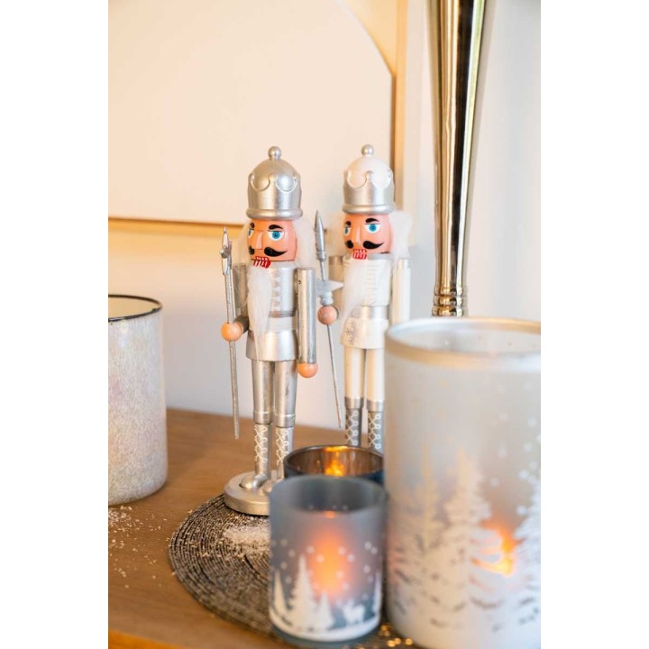 Two silver nutcracker figurines stand beside elegant candles on a warm wooden surface.