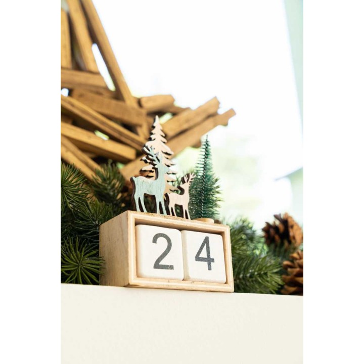 A wooden block calendar with a nature scene of trees and deer, decorated with pine garlands.