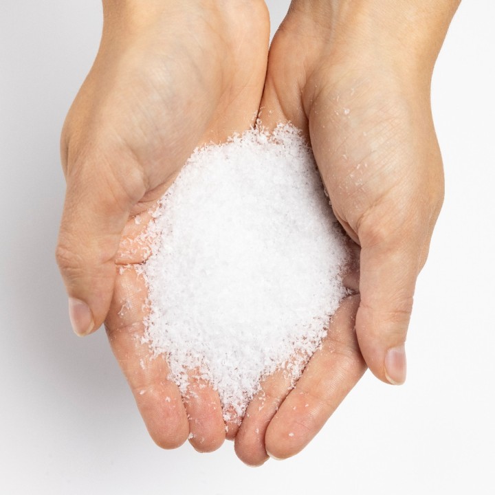 Twee handen houden een bergje witte, kristallijne zout op een lichte achtergrond.