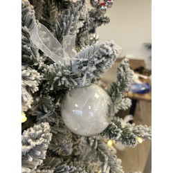 A frosted glass ornament hangs from a snowy evergreen tree, tied with a delicate ribbon.