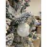 A frosted glass ornament hangs from a snowy evergreen tree, tied with a delicate ribbon.