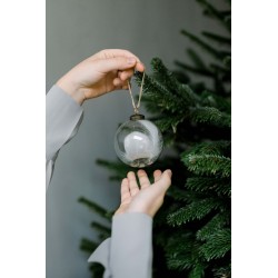 A hand holds a clear glass ornament above another hand, set against a lush green Christmas tree.