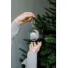 A hand holds a clear glass ornament above another hand, set against a lush green Christmas tree.