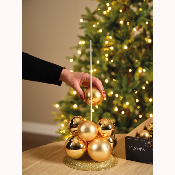 A hand places a shiny gold ornament on a festive display beside a decorated tree.