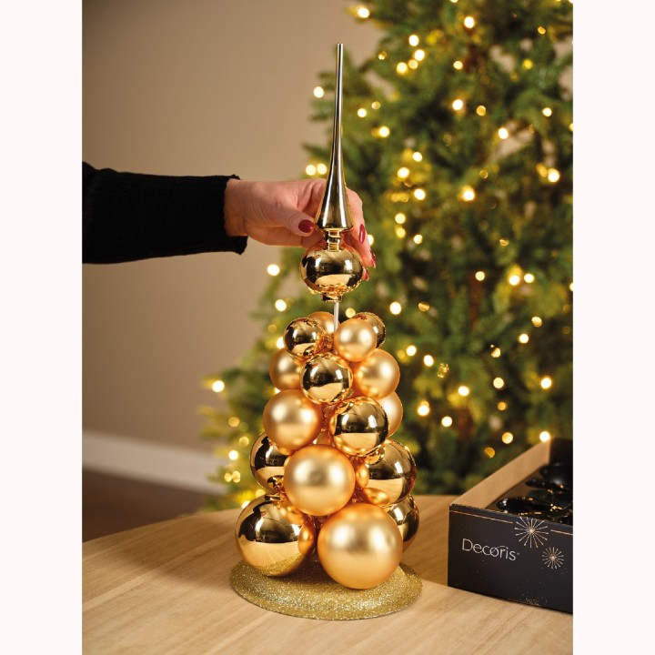 A hand places a shiny gold ornament atop a stacked arrangement of golden spheres.