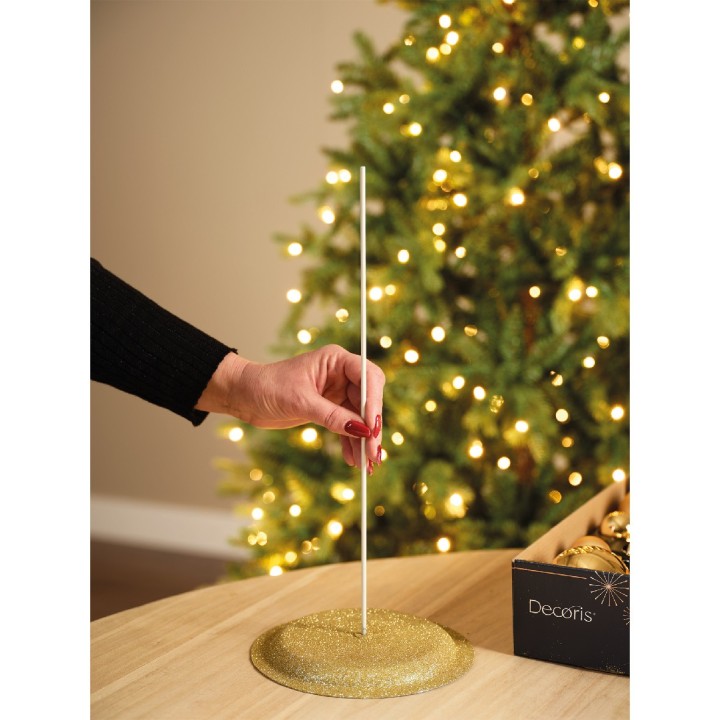 A hand inserts a thin white pole into a glittery gold base near a softly lit Christmas tree.