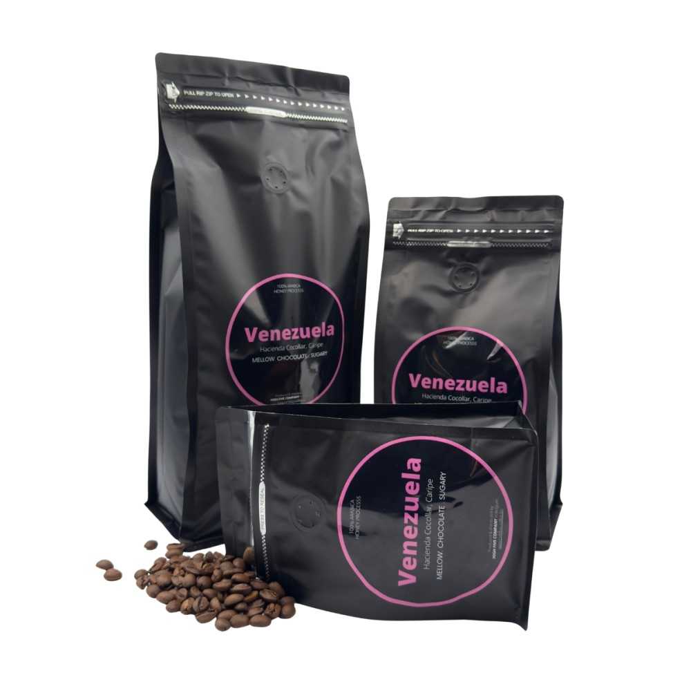 Three black coffee bags with pink labels, accompanied by scattered coffee beans.